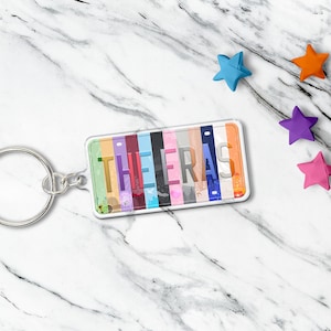 May include: A rectangular keychain with the word "THE ERAS" printed on it. The keychain has a silver key ring and a clear plastic cover. The background of the word is a colorful design. Several star-shaped decorations are scattered around the keychain.