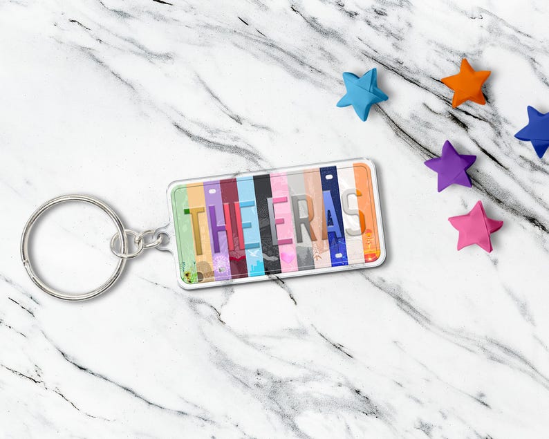 May include: A rectangular keychain with the word "THE ERAS" printed on it. The keychain has a silver key ring and a clear plastic cover. The background of the word is a colorful design. Several star-shaped decorations are scattered around the keychain.