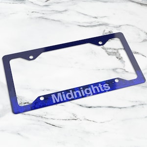 May include: A rectangular license plate frame featuring a dark blue, starry night sky pattern with a subtle gradient effect. The word Midnights is printed in a light blue, sans-serif font across the bottom horizontal section of the frame.
