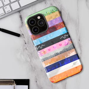 Puede incluir: Una funda de teléfono colorida con un diseño a rayas. La funda presenta varios patrones, incluyendo mariposas, corazones y paisajes, en tonos de rosa, azul, naranja y verde. La funda del teléfono está sobre una superficie blanca junto a un teclado y un bolígrafo.
