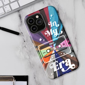 May include: A colorful phone case with the words "In My Era" and a cassette tape design. The case features a vibrant mix of colors, including blue, red, purple, and orange. The phone case is on a white surface with a keyboard and pen.
