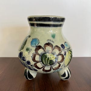 Pode incluir: Um pequeno pote de cerâmica branca com três pernas e um desenho floral. O pote é decorado com flores azuis, verdes, amarelas e marrons.