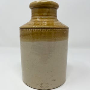 Puede incluir: Un tarro de gres vintage con un esmalte marrón en la parte superior y un esmalte blanco en la parte inferior. El tarro tiene un pequeño sello circular en la parte inferior.