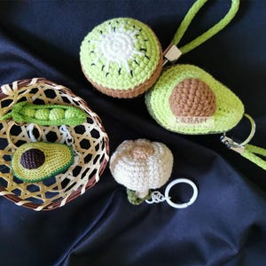 May include: A collection of handmade crochet keychains in the shape of fruits and vegetables. Includes a kiwi, avocado, garlic, and a pea pod with peas. The items are in a woven basket and on a dark background.