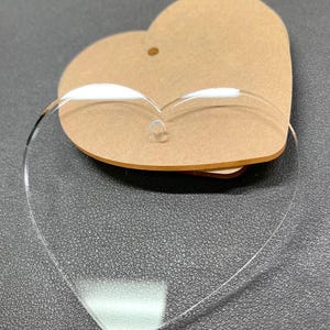 May include: A clear acrylic heart-shaped ornament with a hole at the top for hanging. The ornament is on top of a brown cardboard heart-shaped blank.