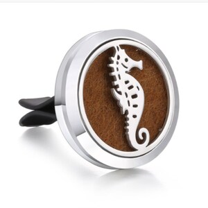 May include: A silver-colored car air freshener with a seahorse design. The seahorse is white and sits on a brown, circular pad. The air freshener has a silver border and a black clip for attachment.