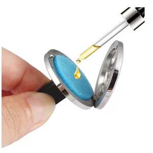 May include: A silver car air freshener with a blue felt pad. A dropper bottle is adding essential oil to the pad.