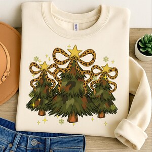 Può includere: Felpa color crema con tre alberi di Natale con motivo mimetico. Ogni albero è adornato con una stella dorata e un fiocco leopardato. Stelle dorate e fiocchi di neve sono sparsi intorno agli alberi.