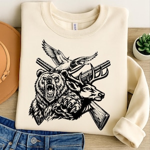 Pode incluir: Camisola creme com um gráfico a preto e branco de um urso, veado e pato com rifles cruzados. O design tem um tema de caça. A camisola está dobrada sobre uma superfície de madeira.