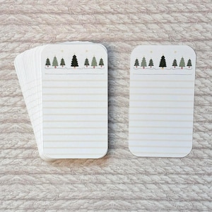 May include: A stack of rectangular, lined notecards with rounded corners. Each card features a row of green and black Christmas trees at the top, with a string of red lights. The cards have a cream-colored background with horizontal lines.