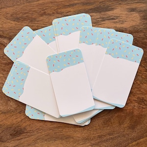 May include: A collection of rectangular paper cards featuring a light blue top section decorated with small pink and yellow sprinkles, transitioning into a solid white bottom section, arranged in a fanned-out overlapping display.