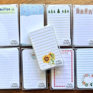 May include: A collection of nine rectangular mini notes pads in clear plastic cases. Designs include a butter label, blue sprinkles, pine trees, gingerbread houses, lined paper, a sunflower, a tea cup, red dots, and a holiday wreath.