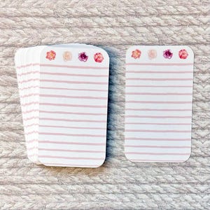 May include: A stack of white notecards with pink horizontal stripes and floral accents. The cards have rounded corners and are arranged on a textured surface. The floral accents include pink, red, and peach-colored flowers.