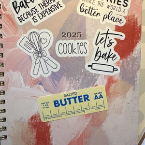 May include: A spiral-bound notebook with a colorful abstract design. The notebook is decorated with stickers that say "Baking because therapy is expensive", "Cookies make the world a better place", "Let's bake", and "Cookies 2025". A butter stick is also visible.