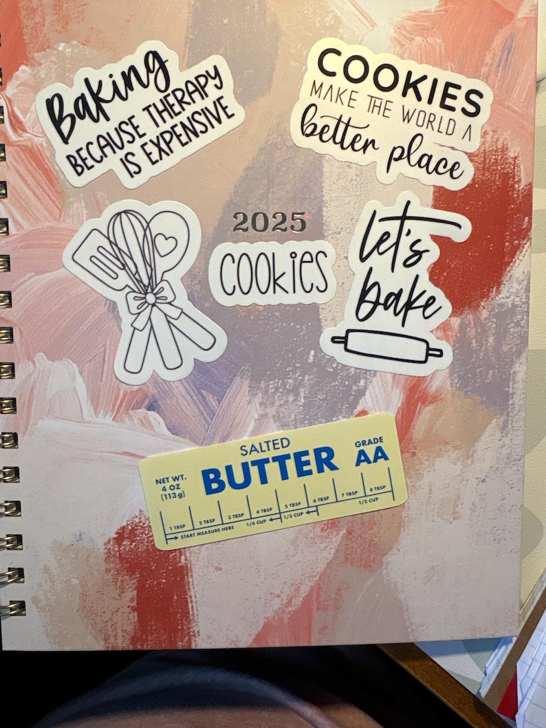 May include: A spiral-bound notebook with a colorful abstract design. The notebook is decorated with stickers that say "Baking because therapy is expensive", "Cookies make the world a better place", "Let's bake", and "Cookies 2025". A butter stick is also visible.