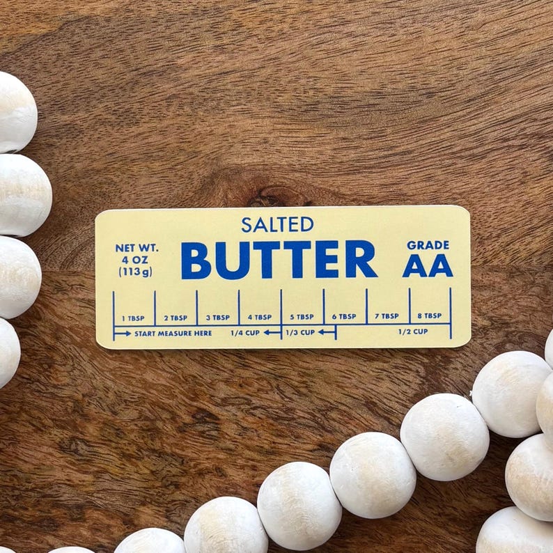 May include: A rectangular, yellow butter measuring guide with the word "BUTTER" in large blue letters. The guide includes measurements in tablespoons and cups, with a net weight of 4 oz (113g). The background is a wooden surface.