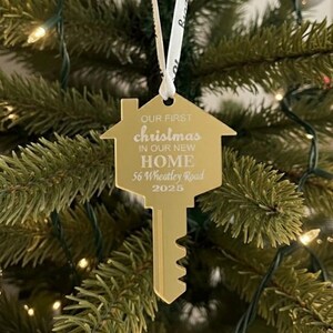 May include: A gold-colored Christmas ornament in the shape of a house key. The ornament has the text "OUR FIRST christmas IN OUR NEW HOME 56 Wheatley Road 2025" in white. The ornament hangs from a white ribbon on a Christmas tree.