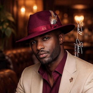 May include: A burgundy felt fedora hat with a matching ribbon band, the initials JB, and a small feather accent. The hat is paired with a burgundy button-down shirt, a beige blazer, and a matching burgundy pocket square with a gold leaf pin.