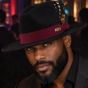 May include: A black fedora hat featuring a wide brim and a deep burgundy ribbon band. The band is accented with the gold-coloured letters R|H and a cluster of iridescent, multi-coloured feathers tucked into the side of the hat crown.