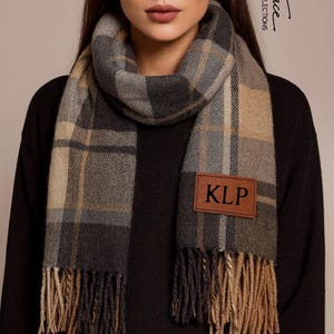 以下が含まれることがあります： グレー、タン、黒のチェック柄のスカーフ。茶色の革のパッチに「KLP」と書かれています。スカーフはフリンジエッジで、首に巻かれています。「Small Space Collections」の文字が背景に見えます。