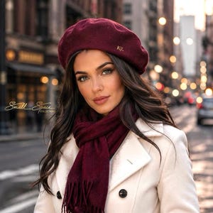 May include: A deep burgundy beret with the gold embroidered letters KJL, a matching burgundy scarf with fringe ends, and matching burgundy gloves. The set is worn with a white double-breasted coat featuring black buttons. The text Small Space Collections is visible on the left.