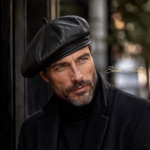 Puede incluir: Una boina de cuero negro con una corona estructurada y una pequeña lengüeta en la parte superior, usada con un suéter de cuello alto negro y un abrigo negro. El texto Small Space es visible en letras doradas a la derecha.