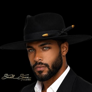 Puede incluir: Un sombrero fedora negro con ala ancha y una cinta negra adornada con dos detalles metálicos en tono dorado en el lateral. El texto Small Space COLLECTIONS es visible en letra blanca en el lado izquierdo de la imagen.