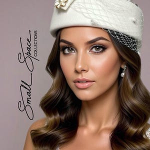 May include: A cream-coloured pillbox hat with a decorative flower and netting. The hat is part of the Small Space Collections. The woman in the image has brown wavy hair and is wearing pearl earrings.