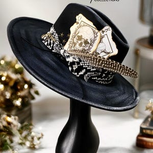 Puede incluir: Un sombrero fedora negro con una banda de estampado de piel de serpiente en blanco y negro, una pluma moteada en marrón y blanco, y dos cartas de juego con un cráneo y un cráneo de toro. El texto Small Space COLLECTIONS se ve arriba.