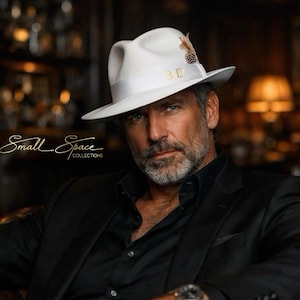 May include: A white fedora hat with a wide white ribbon band featuring the gold letters BD and a small decorative feather accent on the side. The hat has a structured crown and a flat brim, presented in a clean, solid white felt material.