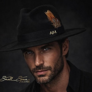 May include: A black fedora hat featuring a wide brim, a black ribbon band with the gold text A|M, and a brown and black patterned feather accent. The text Small Space COLLECTIONS is visible in gold on the left side of the image.