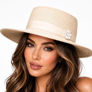 May include: A beige straw fedora hat with a wide brim and a matching beige ribbon band. A small white flower-shaped embellishment is attached to the side of the ribbon. The text Small Space COLLECTIONS is printed in gold at the bottom.