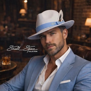 May include: A light blue suit with a white dress shirt and a fedora hat with a blue band and feather detail. The hat has the initials "JLD" on the band. The image also shows a silver wristwatch. The text "Small Space Collections" is visible.