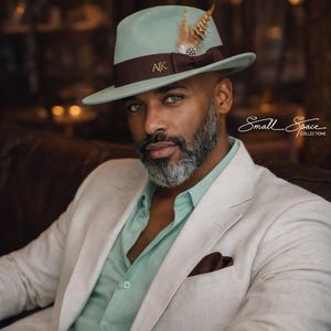 Puede incluir: Un sombrero fedora verde claro con una banda marrón y detalles de plumas. El sombrero tiene las iniciales "AK" en la banda. La persona lleva una camisa abotonada verde claro y una blazer color crema. La imagen es de Small Space Collections.