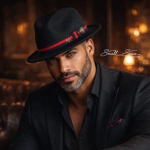 Puede incluir: Un sombrero fedora negro con una banda roja y detalles dorados. El sombrero se usa con un traje negro y un reloj de pulsera dorado. Un vaso de líquido ámbar se encuentra sobre una superficie de madera.