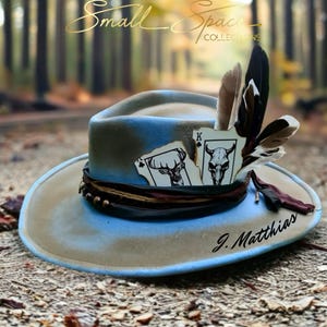May include: A tan and blue felt hat featuring a dark band, a cluster of brown and white feathers, and two playing cards depicting a deer head and a skull. The text Small Space COLLECTIONS and J. Matthias are visible on the hat and background.