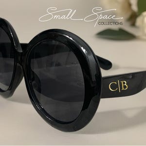 May include: Oversized black sunglasses with round frames and dark lenses. The temples feature the gold-colored letters C|B. Above the sunglasses, the text Small Space COLLECTIONS is visible in a white script and sans-serif font combination.