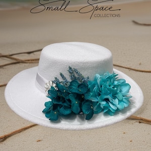 Puede incluir: Un sombrero blanco de ala ancha, adornado con detalles florales turquesa y azules. El sombrero presenta una cinta blanca y el texto "Small Space Collections" en una escritura elegante.