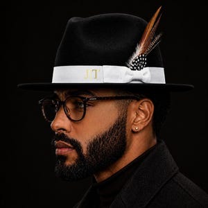 May include: A black fedora hat featuring a wide white ribbon band with the gold embroidered letters JT. The hat is adorned with a cluster of brown, black, and white patterned feathers tucked into the side of the white ribbon band.