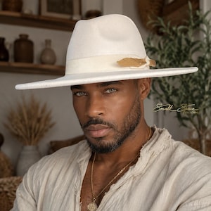 Puede incluir: Un sombrero fedora de color blanco roto con ala ancha, una cinta a juego y un pequeño adorno de pluma dorada en el lateral. El sombrero tiene una copa estructurada y un ala plana. El texto Small Space Collections es visible a la derecha.