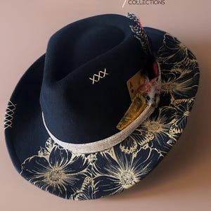 Puede incluir: Un sombrero fedora azul marino con un diseño floral en el ala y una banda plateada. El sombrero presenta una pluma y una carta de juego. El texto "Small Space Collections" es visible.