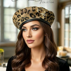 May include: A tan and black leopard print beret featuring a black leather trim and a gold-toned bee brooch with clear crystals. The text Small Space COLLECTIONS is visible at the top of the image above the beret.