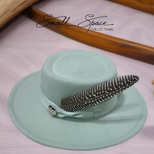 May include: A light blue fedora hat with a decorative feather and a ribbon band. The hat features a small, round, silver-colored button on the ribbon. The text "Small Space Collections" is written in cursive on the top of the image.