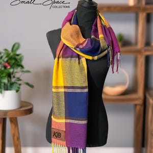 Puede incluir: Una bufanda a cuadros colorida en tonos amarillos, morados, azules y naranjas se muestra en un maniquí negro. La bufanda tiene un borde con flecos y un parche de cuero con las iniciales "K/B". El texto "Small Space Collections" es visible en la esquina superior izquierda.