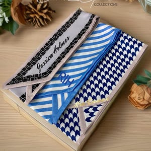 May include: A wooden box containing three silk scarves with various patterns. One scarf is light gray with black geometric designs and the name "Jessica Holmes". Another is blue and white striped, and the third is navy blue with white arrow patterns.