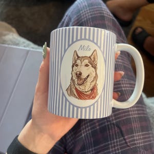 Może przedstawiać: Biały ceramiczny kubek w jasnoniebieskie i białe pionowe paski. Na środku znajduje się owalna ramka z ilustracją brązowo-białego psa rasy husky w czerwonej bandanie, z imieniem Milo napisanym nad głową psa.