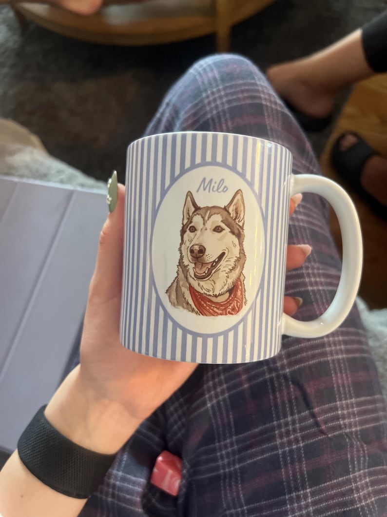 Może przedstawiać: Biały ceramiczny kubek w jasnoniebieskie i białe pionowe paski. Na środku znajduje się owalna ramka z ilustracją brązowo-białego psa rasy husky w czerwonej bandanie, z imieniem Milo napisanym nad głową psa.