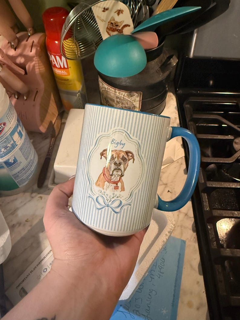Może przedstawiać: Ceramiczny kubek w jasnoniebieskie i białe pionowe paski. Z przodu znajduje się ozdobna ramka z ilustracją brązowego psa rasy bokser w czerwonej obroży, z napisanym niebieską czcionką imieniem Bigby nad głową psa.