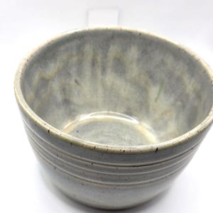 May include: A handmade ceramic bowl with a speckled, light gray glaze. The bowl has a rounded shape with a slightly flared rim and horizontal grooves around the exterior. The interior is a lighter shade of gray.