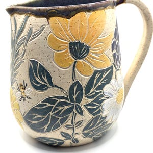 May include: A ceramic pitcher with a cream-colored base, featuring embossed floral and leaf designs in yellow, white, and teal. The handle and rim are a speckled cream color, with a dark brown rim. A bee is also embossed on the side.